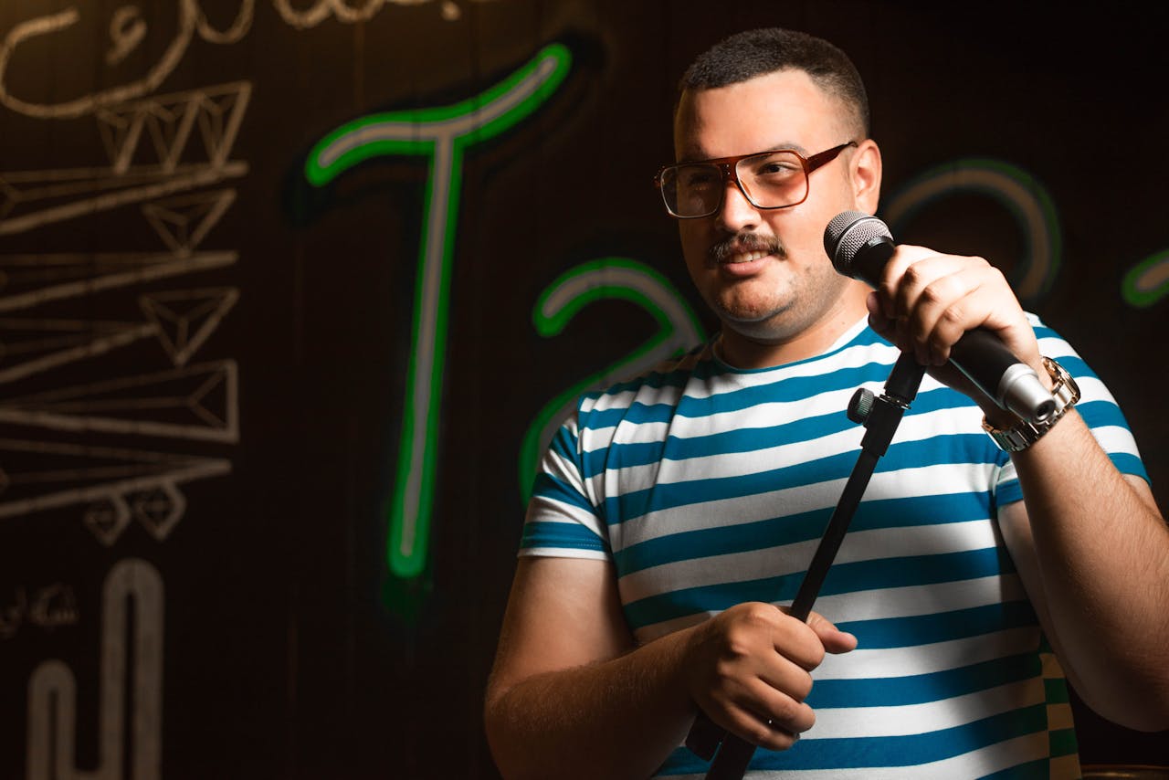 Man holding a microphone doing stand-up comedy in casual striped shirt on stage.