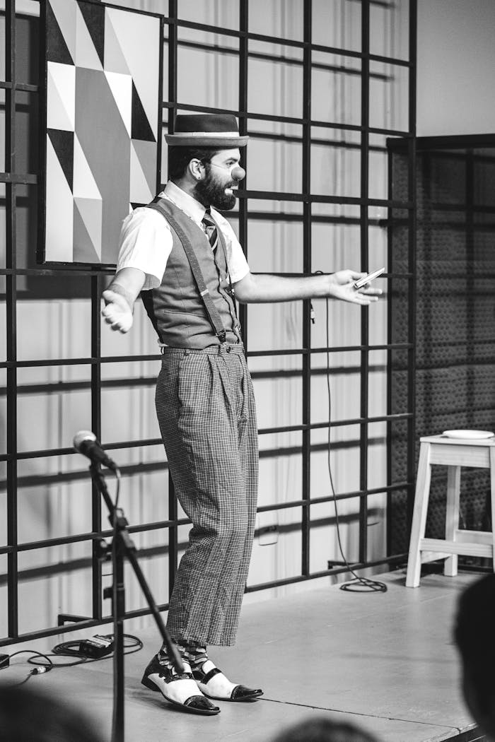 A black and white image of a male comedian performing on stage, dressed in vintage attire.